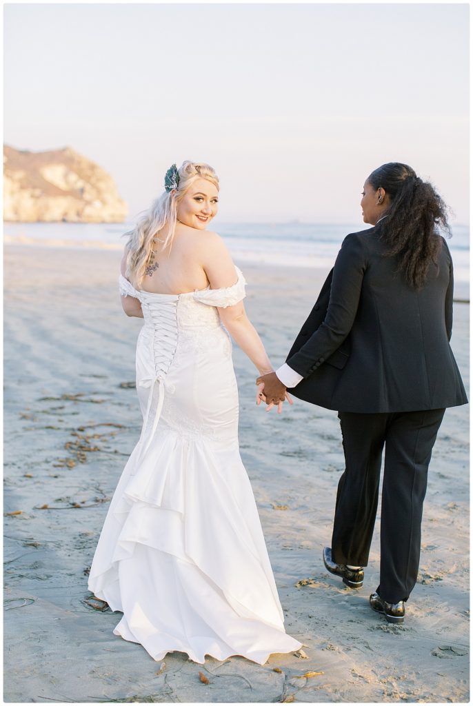 2 brides walking on the beach but one bride is looking back at the photographer