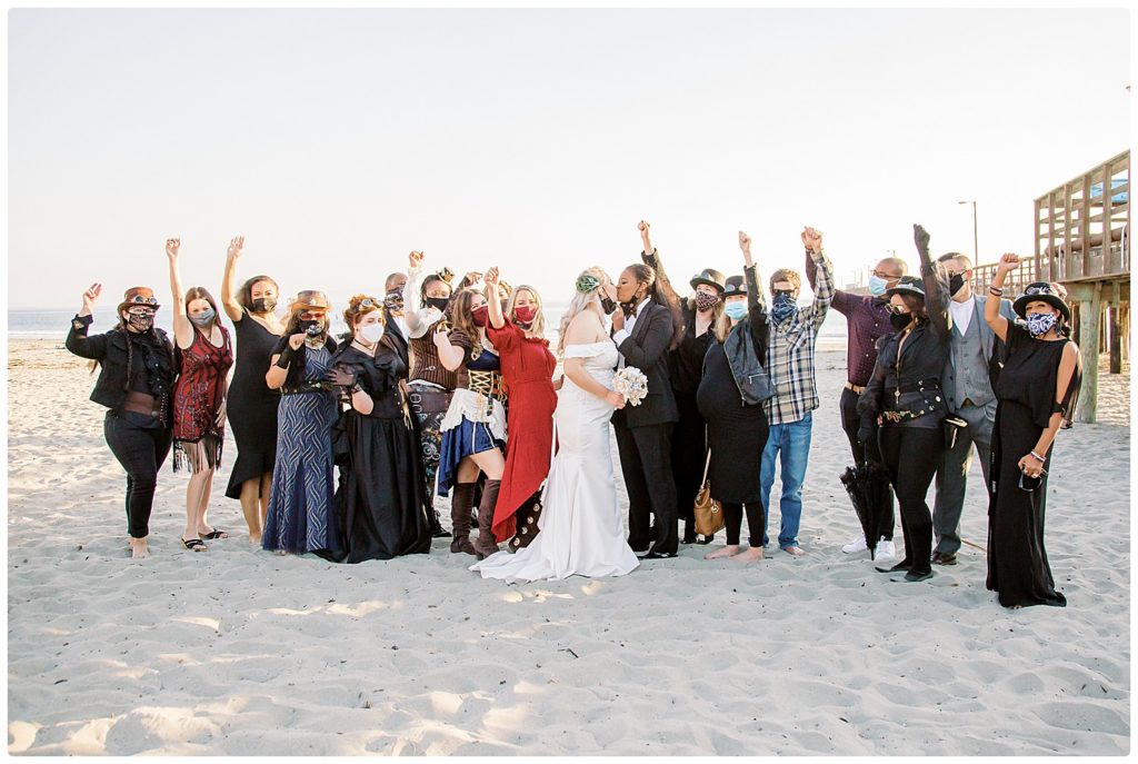Wedding guests cheering while 2 brides kiss during steampunk beach wedding in California