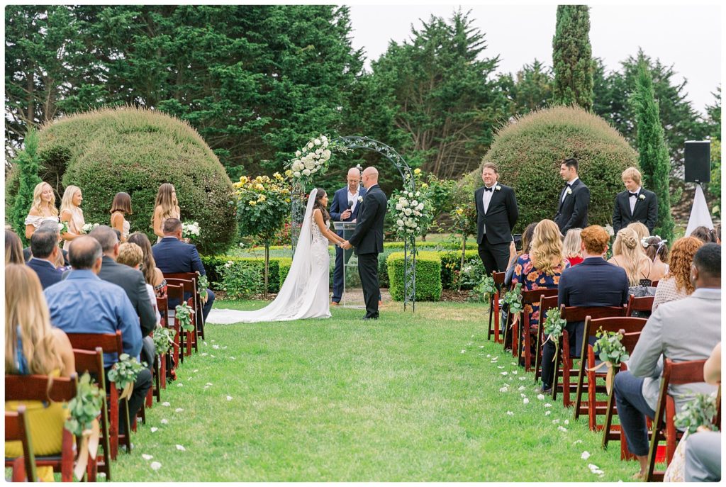 Wedding party with bride and groom holding hands and staring into each other's eyes at their garden wedding at Heritage Estates