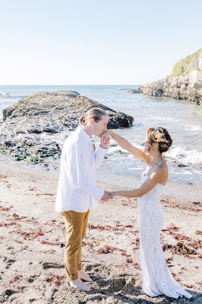 groom kissing the bride's hand while on the beach