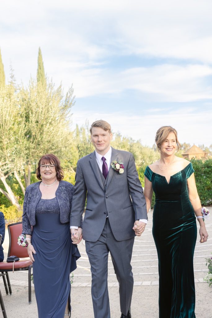 groom walks down the aisle with his mom and bride's mom